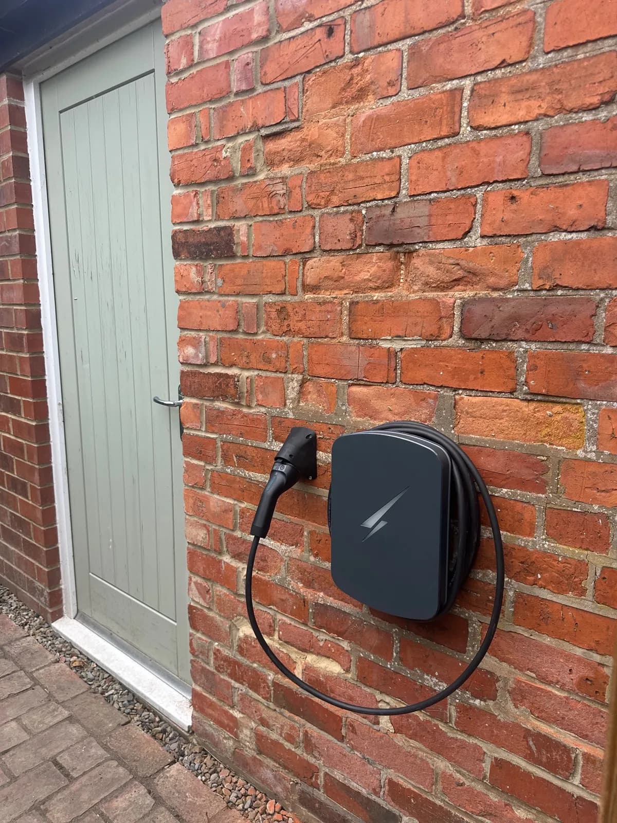 Graphite grey home EV charger with its cable coiled, fixed to an old reclaimed red brick wall beside a garden door