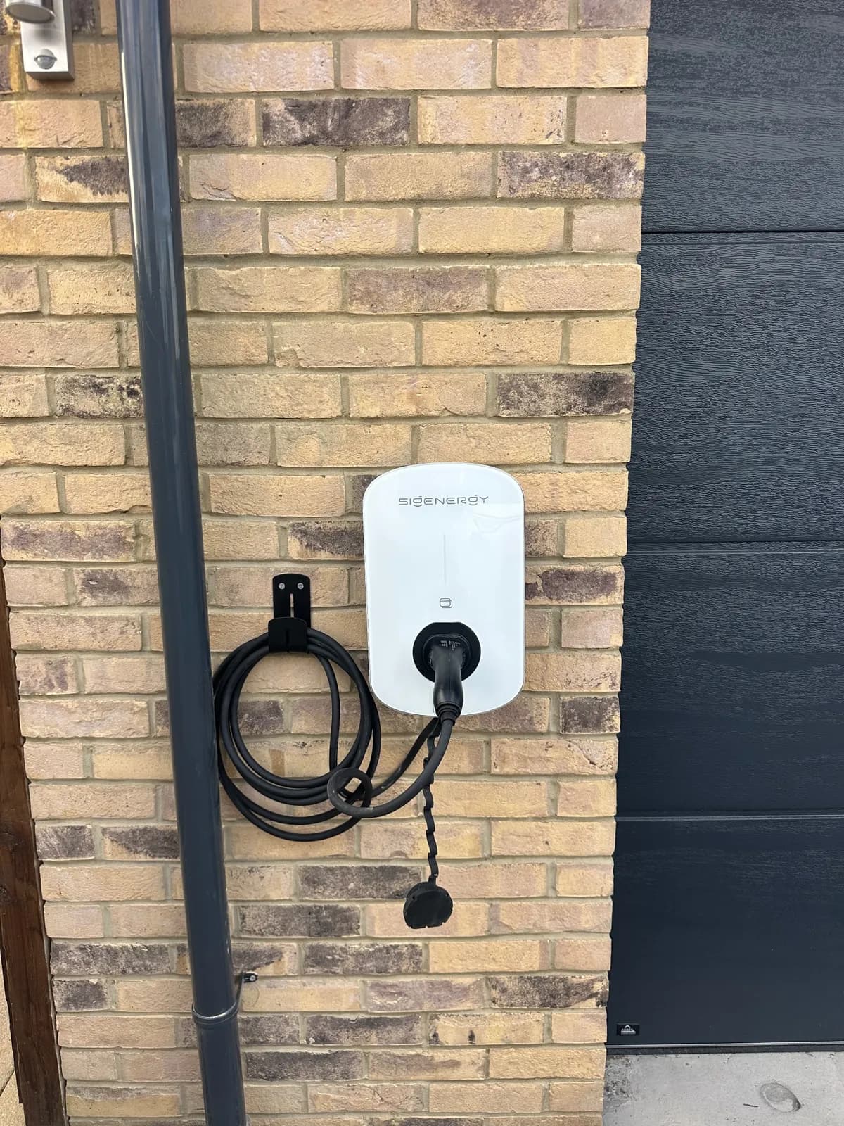 Sigenergy EV charger with a cable hook, fitted on buff brick next to a grey garage door